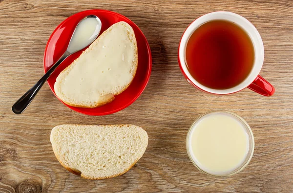 Sandwich with condensed milk, teaspoon in saucer, red cup with tea, slice of bread, glass bowl with milk on wooden table. Top view