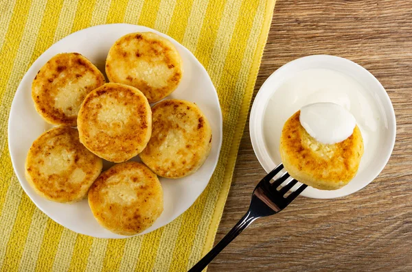 Homemade small cottage cheese pancakes in white plate on yellow striped napkin, pancake strung on fork above bowl with sour cream on wooden table. Top view