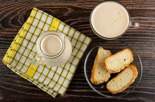 Pitcher with milk on checkered napkin, cup with fermented baked milk, pieces of sprinkled horn-shaped buns in transparent glass saucer on dark wooden table. Top view