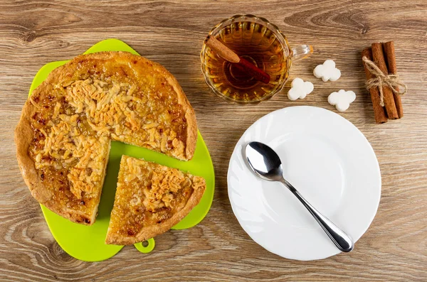 Pieces of sweet lemon pie on green cutting board, cup of tea with cinnamon stick, sugar cubes, bunch of cinnamon sticks, teaspoon on white plate on wooden table. Top view