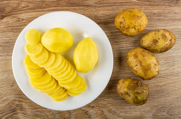 Raw peeled potatoes and slices of potato in white plate, unpeeled potatoes on wooden table. Top view