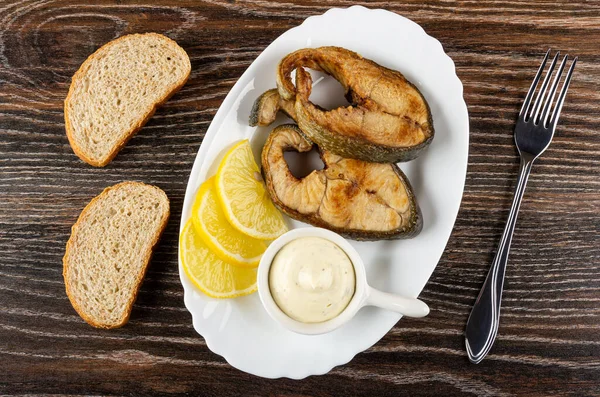 Pieces of bread, slices of lemon, sauce boat with mayonnaise, fried steak from salmon in white dish, fork on dark wooden table. Top view