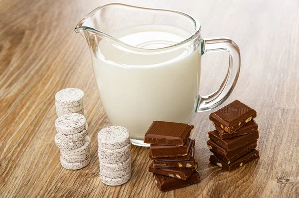 Pitcher with milk, stacks of candy from powdered milk and chocolate in shape tablets, stacks of dairy chocolate on wooden table