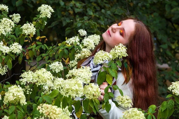 Frau mit roten Haaren und hellem Make-up in der Nähe von Blumen in der Natur — Stockfoto