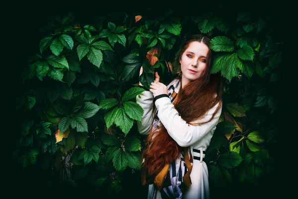 Donna con lunghi capelli rossi e trucco luminoso vicino alle foglie verdi — Foto Stock