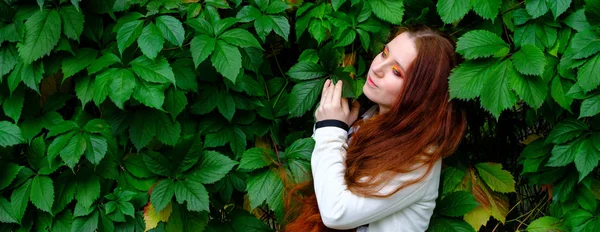 Женщина с длинными рыжими волосами и ярким макияжем рядом с зелеными листьями — стоковое фото