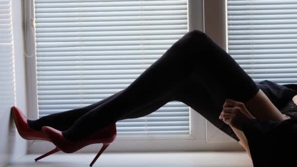 Hosszú női lábak fekete harisnyában és piros magas sarkú cipőben az ablakpárkányon redőnyökkel — Stock videók