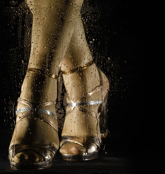 female feet in high heeled sandals behind glass with water drops on black background