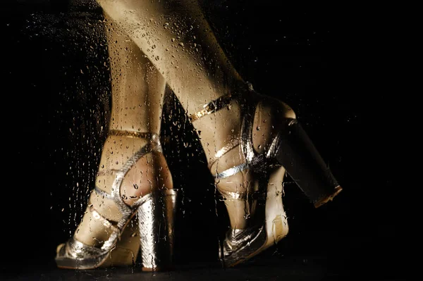 female feet in high-heeled sandals behind glass with water drops on black backgroun