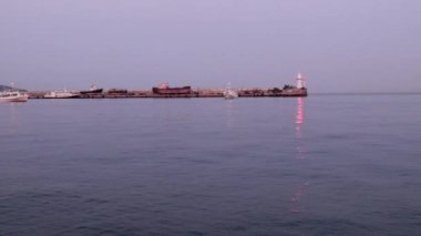 Bir ticaret gemisinin hareketi ve limanın su alanındaki iskelede titreşen bir deniz feneri.