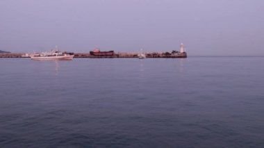 Bir ticaret gemisinin hareketi ve limanın su alanındaki iskelede titreşen bir deniz feneri.
