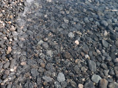 Bir çakıl taşı plajının yüzeyi ve berrak deniz suyu dalgaları.