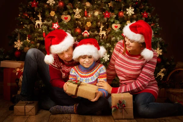Χριστουγεννιάτικο οικογενειακό ανοιχτό δώρο μπροστά από το χριστουγεννιάτικο δέντρο, ευτυχείς γονείς με παιδί σε Santa Hats — Φωτογραφία Αρχείου