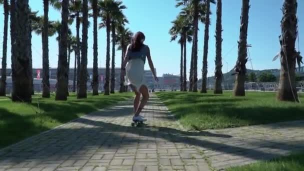 Hipster moderno jovem senhora passeios no skate bordo no beco de palmeiras seu vestido e cabelo são soprados no vento e brilhar brilhante — Vídeo de Stock