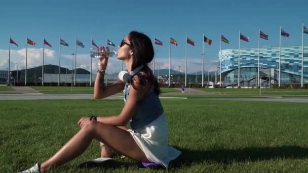 Jeune brune bronzée sportive buvant lentement de l'eau tout en étant assise sur l'herbe verte entourée de bâtiments modernes — Video