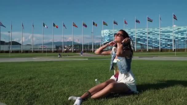 Steet punk brun ung brunette liker sommer mens sitter på det grønne friske gresset omgitt av moderne bygninger – stockvideo
