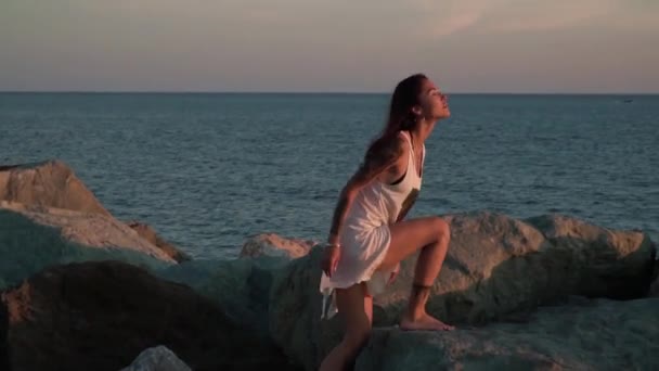 Gadis berambut cokelat memanjat batu laut besar menuju cahaya matahari terbenam sementara gaun putih dan rambutnya tertiup angin — Stok Video
