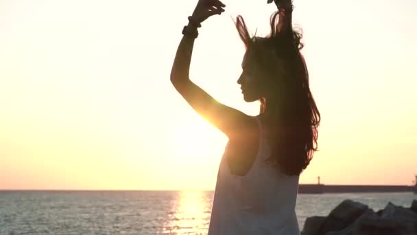 Verführerisches Mädchen mit langen schwarzen Haaren in einer transparenten Tunika, die bei Sonnenuntergang am Strand posiert — Stockvideo
