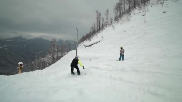 Los paseos en snowboarder se deslizan tentativamente por la nieve fresca en la estación de esquí. Clima nublado y brumoso en la estación de esquí. Paseos en snowboarder rodeados de fascinantes montañas . — Vídeos de Stock