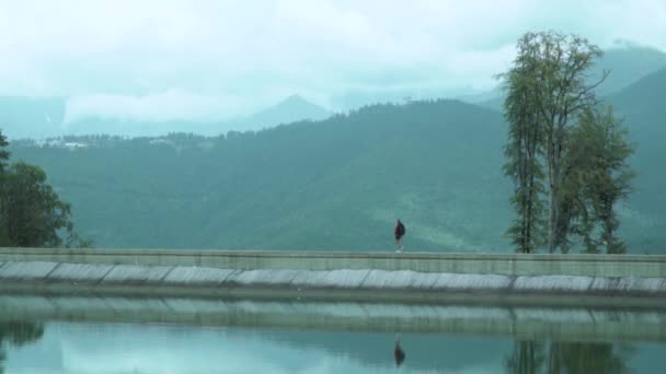 Tiny figure of hiker woman and her reflection at the mirror water surface of mountain lake against distant fog-hided huge peacks — Stock Video