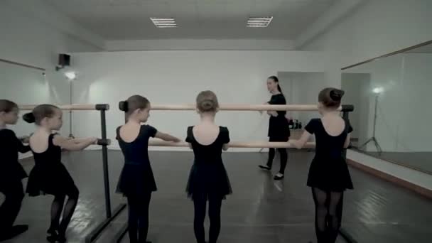 La morena profesora de clases de baile muestra a las pequeñas bailarinas vestidas de maillot negro cómo pararse cerca de la barra de ballet — Vídeo de stock