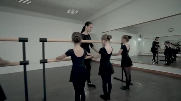 ダンスクラスのブルネットの女性教師は、バレエバレエの近くに立つ方法を黒いレオタードに身を包んだリトルバレリーナに示します — ストック動画