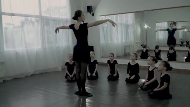 Ballerina stand at front of few littel balerinas who look at her. Adult Ballerina teach littel balerinas how to move slowly and they look at her. Littel balerinas are sitting on the floor and look at — Stock Video