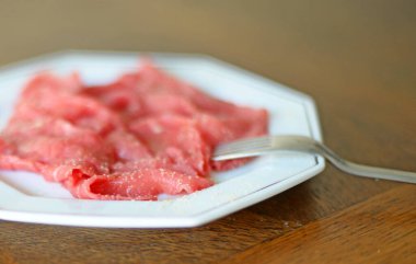 Zeytinyağlı sığır eti carpaccio ve beyaz porselen kaplama beyaz biber üzerinde gümüş astar ve paslanmaz çelik çatal, kahverengi meşe ağacından arka plan. Çok sığ alan derinliği