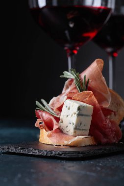 Lezzetli aperatif ve bardak kırmızı şarap. Jambon, mavi peynir ve karanlık bir arka plan üzerinde biberiye ile sandviç. Metniniz için kopya alanı