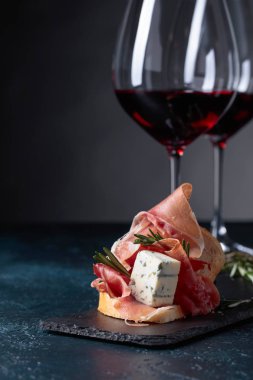 Lezzetli aperatif ve bardak kırmızı şarap. Jambon, mavi peynir ve karanlık bir arka plan üzerinde biberiye ile sandviç. Metniniz için kopya alanı