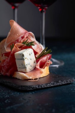Lezzetli aperatif ve bardak kırmızı şarap. Jambon, mavi peynir ve karanlık bir arka plan üzerinde biberiye ile sandviç. 