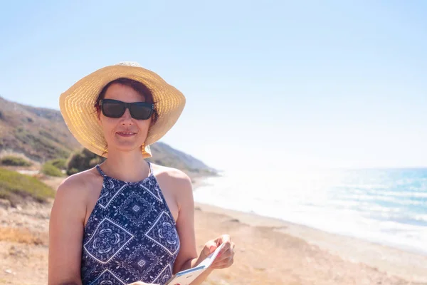 在地中海海岸戴墨镜和帽子的中年妇女 女人用手机看着摄像机 夏日阳光明媚的一天 复制空间 — 图库照片