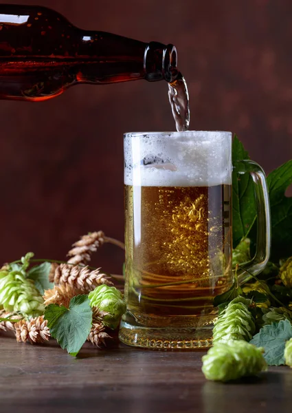 Mug of beer, grain and hops on a old wooden table. Beer is poured into a mug from the bottle. Free space for text.