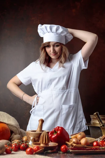 年轻漂亮的女孩在一个厨师制服认为在桌子前与各种厨房用具和蔬菜 — 图库照片