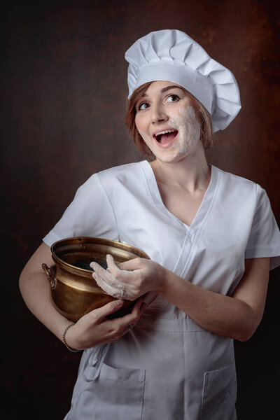 Happy young beautiful girl in a chef uniform with old pan on a brown backgroud. Cheerful woman soiled with flour.