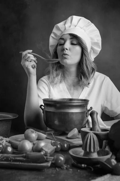 年轻漂亮的女孩在一个厨师制服与老黄铜平底锅和木勺品尝食物 餐桌上的各种厨具和蔬菜 — 图库照片