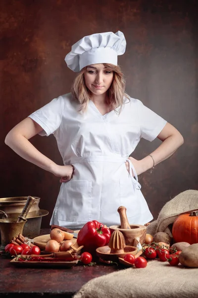 穿着大厨制服的年轻漂亮女孩在餐桌前想着各式各样的厨具和蔬菜 素食主义主题的概念意象 — 图库照片