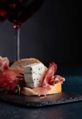 Jambon, mavi peynir ve karanlık bir arka plan üzerinde biberiye ile sandviç. Lezzetli aperatif ve bardak kırmızı şarap. Metniniz için kopya alanı