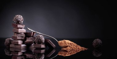 Çikolata şekerleme, çikolata parçaları ve kaşık kakao tozu ile siyah bir yansıtıcı arka plan üzerinde kırık.