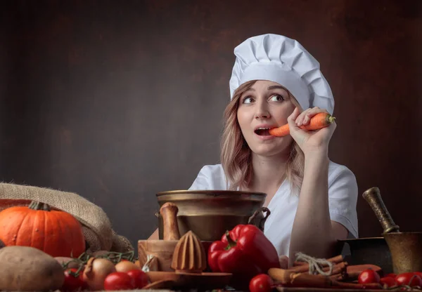 年轻漂亮的女孩在厨师的制服吃胡萝卜 桌子上 不同的厨房用具和蔬菜 素食主义主题的概念意象 — 图库照片