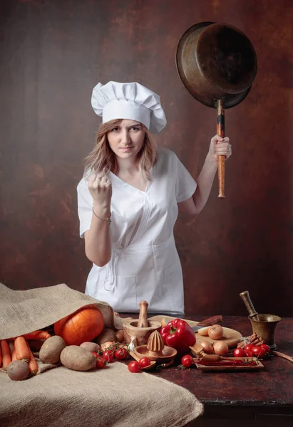 Very angry chef with big frying pan in hand. On the table different kitchen utensils and vegetables.