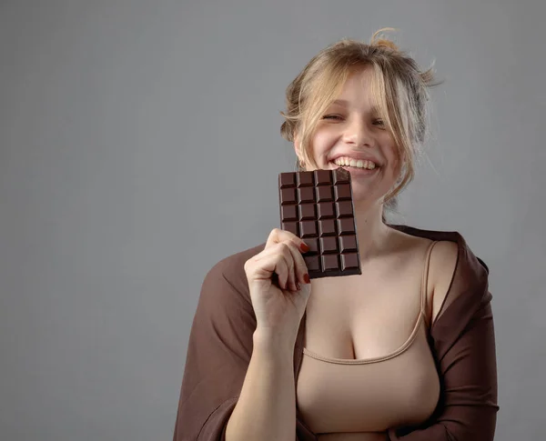 幸せな若い美しい女性のチョコレートを食べるします 灰色の背景に分離されたチョコレートを食べると楽しい自然化粧品で若い金髪 — ストック写真