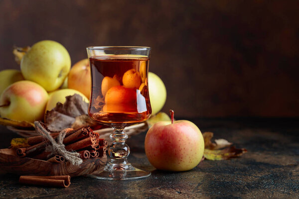 Glass of apple juice or cider with juicy apples and cinnamon sticks on a kitchen table.