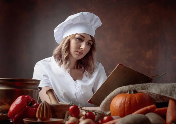 穿着大厨制服的年轻漂亮女孩读了一本老食谱 餐桌上 不同的厨房用具和蔬菜 — 图库照片