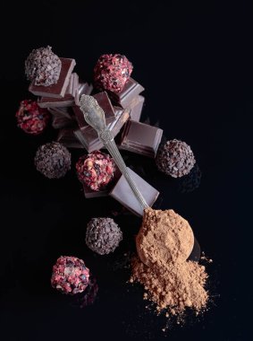 Çikolata şekerleme, çikolata parçaları ve kaşık kakao tozu ile siyah bir yansıtıcı arka plan üzerinde kırık.