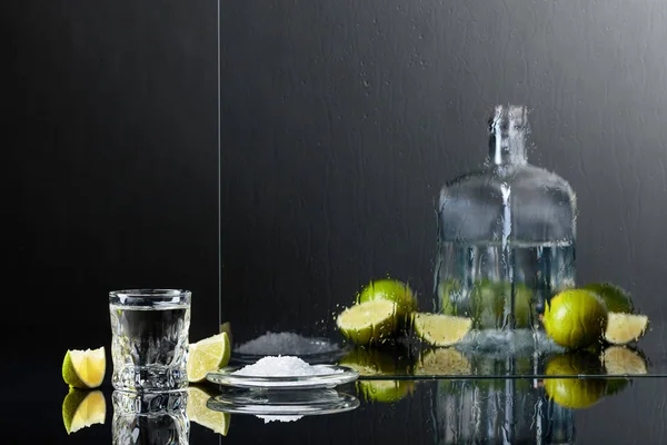 Glass of tequila with lime and salt on the black reflective background. Alcohol in glass before wet glass with water drops.