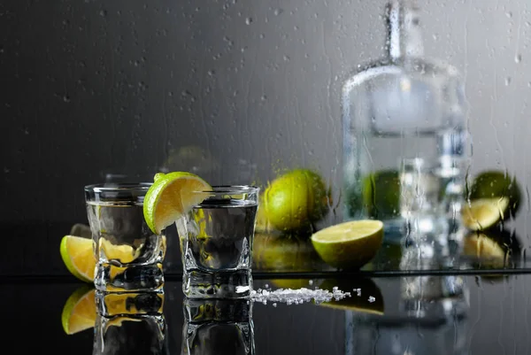 Glasses of tequila with lime and salt on the black reflective background. Alcohol in glasses before wet glass with water drops.