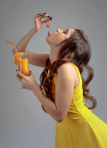 年轻美丽的女孩在一个黄色的衣服与樱桃 手里拿着鸡尾酒龙舌兰日出 用橘子片装饰 — 图库照片