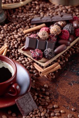 Çeşitli çikolatalar ve bir fincan sade kahve. Tarçın çubuklu tatlılar ve kahve çekirdekleri kahverengi bir masada..