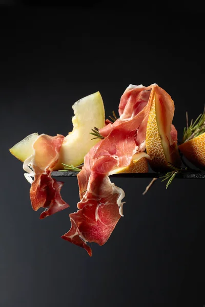 Prosciutto med melon och rosmarin på svart bakgrund. — Stockfoto